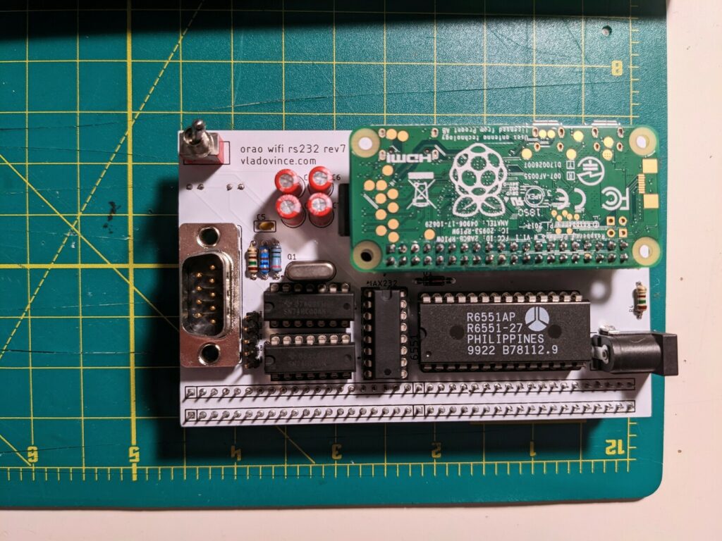 Front of a white PCB board with a number of chips, a green raspberry pi zero, a db9 port and a large SPDT switch. The board says orao WiFi rs232 rev7 in the upper left corner